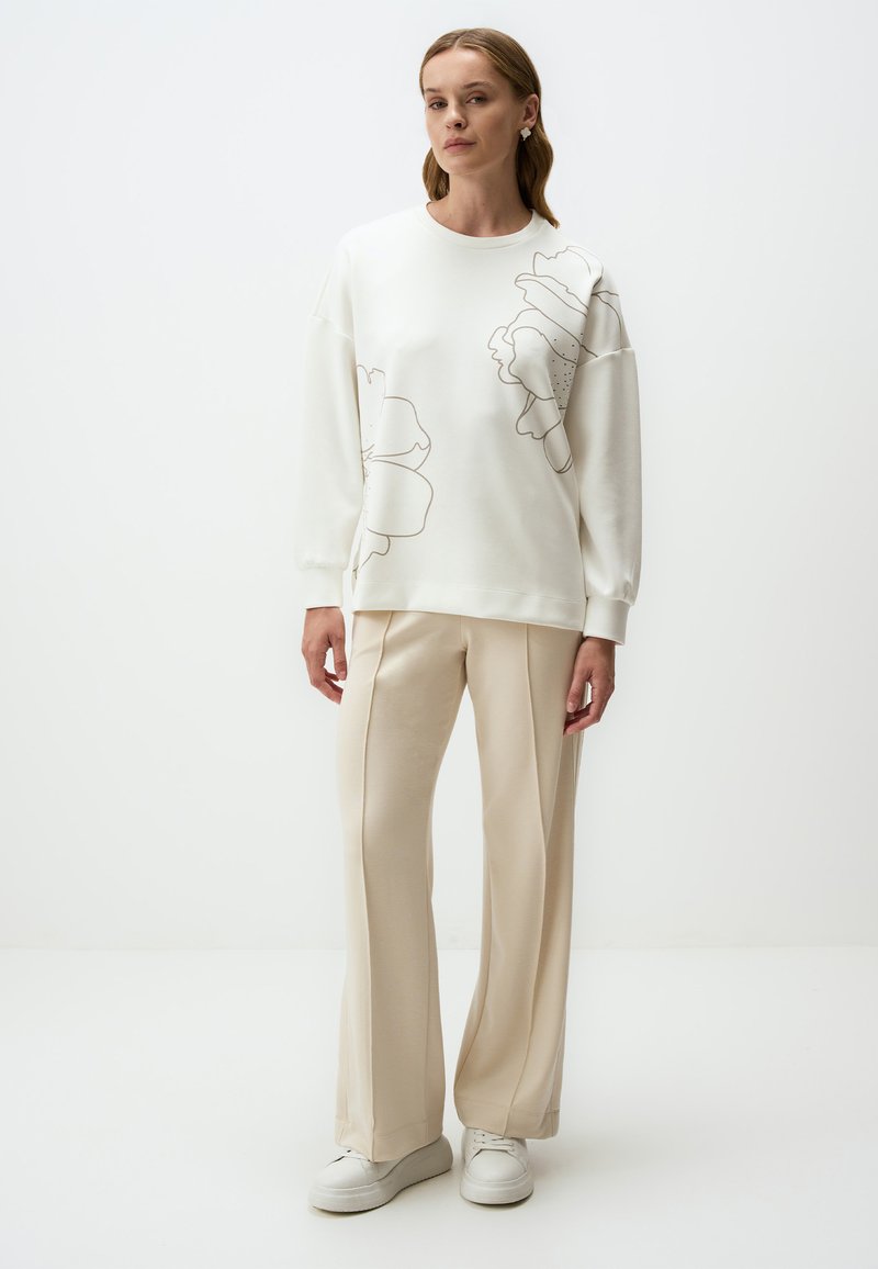 Sweatshirt blanc avec un grand motif floral en contours, associé à un pantalon beige à jambe large. Texture lisse, coupe décontractée et baskets blanches.
