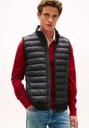 Svart vattert vest med vertikalt quiltet design, glidelås foran og sidelommer, brukt over en rød genser, kombinert med blå jeans.