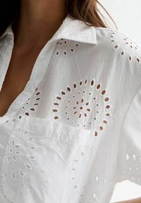 White cotton blouse featuring intricate eyelet embroidery with circular designs, crisp collar, and a relaxed fit with a chest pocket.