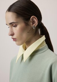 Lichtgroene trui met een brede gele kraag. Glad textuur, aansluitend ontwerp en een zilveren oorbel aan de linkerkant. Minimalistische stijl.