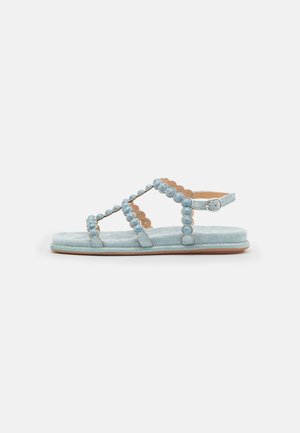 Sandalia de ante azul claro con diseño de tiras cruzadas, detalles redondeados, correa ajustable en el tobillo y suela plana. Textura suave y elegancia sencilla.