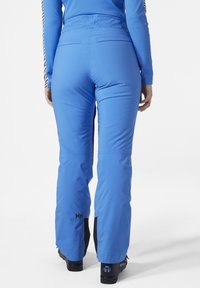 Modré voděodolné lyžařské kalhoty s hladkým povrchem, které mají černé akcenty, pás na střední výšku a logo viditelné na dolní části nohy.