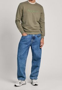 Mężczyzna w oliwkowym swetrze Pepe Jeans, w luźnych niebieskich dżinsach i białych sneakersach, stoi z jedną ręką w kieszeni na jednolitym tle.