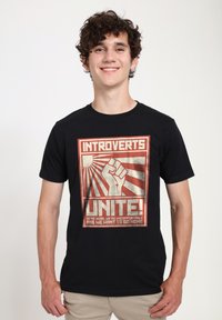 Sort bomulds T-shirt med et grafisk print af en løftet næve, stråler og tekst: "INTROVERTE FORENEN SIG! VI ER HER, VI ER UBEHAGELIGE."