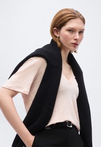 Γυναίκα με κοντά μαλλιά που φοράει ένα ανοιχτόχρωμο T-shirt με V λαιμόκοψη, μαύρο παντελόνι και ένα μαύρο πουλόβερ περασμένο στους ώμους, με χρυσούς κρίκους στα αυτιά.