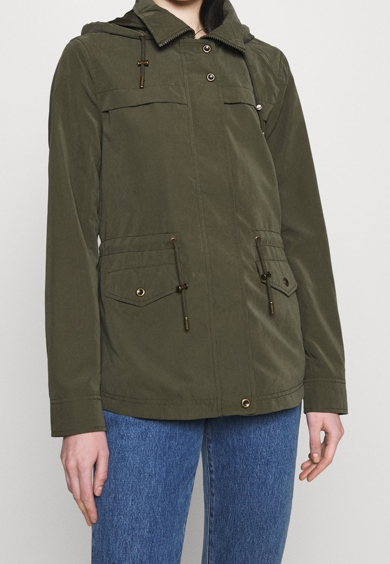 Personne portant une veste à capuche vert olive avec poches avant et cordons de serrage, associée à un jean en denim bleu.