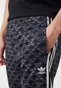 Czarne spodnie sportowe z szarym wzorem liści, elastycznym paskiem w talii, bocznymi kieszeniami oraz białymi pionowymi paskami po bokach i logo Adidas.