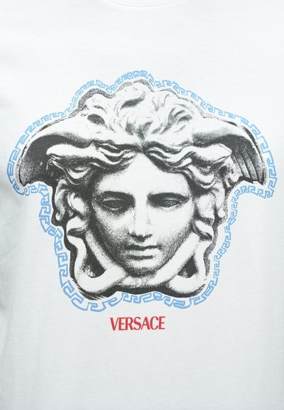 Λευκό μπλουζάκι με εκτύπωση μιας ασπρόμαυρης Μέδουσας περιτριγυρισμένης από ένα μπλε μοτίβο ελληνικού κλειδιού, με την ένδειξη "VERSACE" τυπωμένη με κόκκινα γράμματα από κάτω.