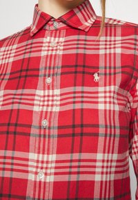 Röd rutigt skjorta i tyg med vita och svarta accenter. Har krage med knappar och en liten broderad logotyp på bröstet.