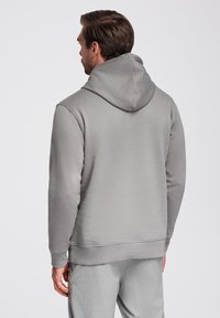 Šedá mikina s kapucí, měkká tkanina, volný střih, žebrované manžety a pas. Ševový detail na kapuci, hladký zadní díl, bez grafiky nebo zdobení.