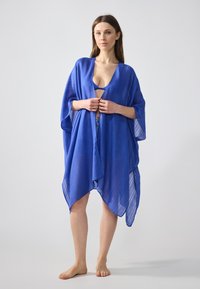 Blå, lätt kimono-liknande överdrag i texturerad tyg, med breda ärmar och asymmetrisk fåll. Ingen stängning finns.
