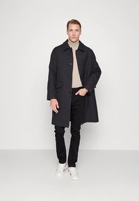 Dunkler Wollmantel mit Kragen, Frontknöpfen und zwei Seitentaschen, getragen über einem beigen Rollkragenpullover, kombiniert mit schwarzen Hosen und weißen Turnschuhen.