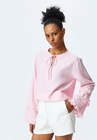 Maglietta a maniche lunghe rosa chiaro con scollo rotondo, dettaglio di nodo frontale e accenti di polsini arricciati, abbinata a pantaloni corti bianchi. Tessuto morbido e testurizzato.