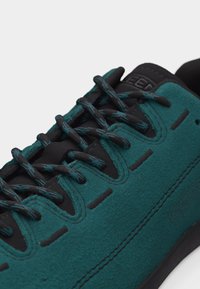 Κοντινό πλάνο αθλητικού παπουτσιού σε τυρκουάζ και μαύρο χρώμα με κορδόνια με σχέδιο και ορατή ραφή, που αναδεικνύει το πάνω μέρος και την περιοχή των κορδονιών.