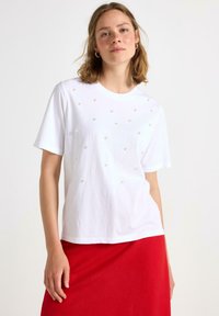 Hvid bomulds T-shirt med korte ærmer, prydet med forhøjede perle-lignende dekorationer arrangeret i et gittermønster over fronten.