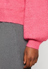 Main noire, détendue sur le côté, portant un pull en tricot rose avec des manches bouffantes et une jupe ou un pantalon gris texturé.