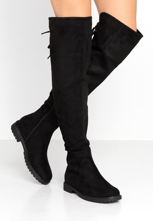 Mustat polven yli ulottuvat tasaiset saappaat, joissa on sivuvetoketju ja uritettu pohja. Saappaita käyttää henkilö, joka istuu jalat ristissä valkoista taustaa vasten.