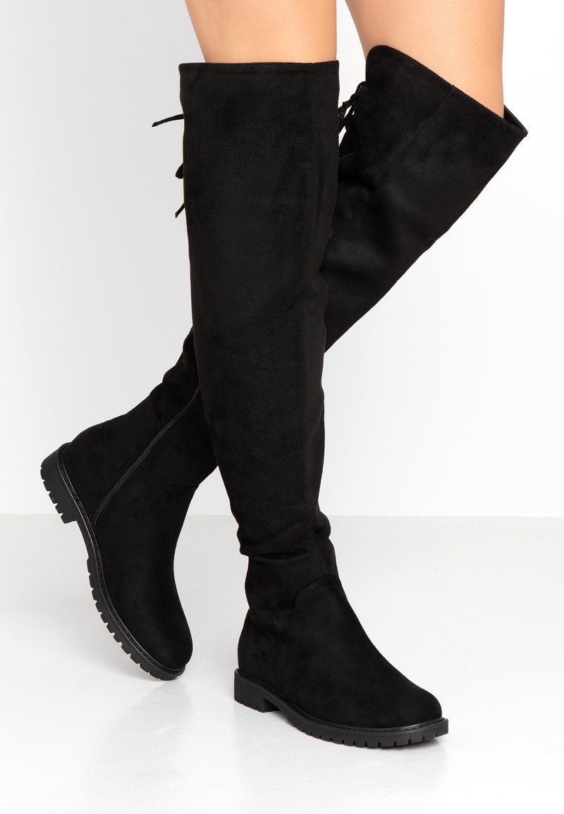 Bottes plates noires montantes au-dessus du genou avec fermeture éclair latérale et semelle crantée, portées par une personne aux jambes croisées sur un fond blanc.