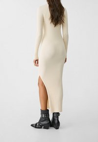 Geripptes, figurbetontes Kleid in Beige mit langen Ärmeln und hohem Schlitz. Kombiniert mit schwarzen Leder-Stiefeletten mit Metalldetails.