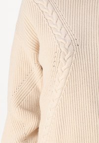 Pull en tricot beige avec des motifs texturés variés, y compris des détails en torsades et des côtes sur les manches et le corps.