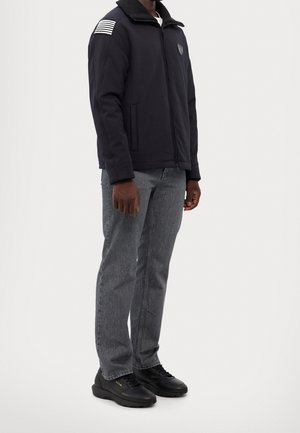 Mężczyzna w czarnej kurtce zapinanej na zamek z paskiem w paski na rękawie, szarych dżinsach i czarnych tenisówkach stoi na tle jednolitego tła.