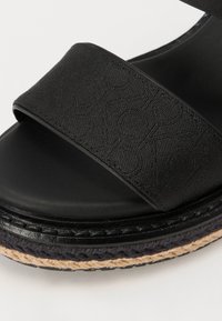 Sandalias negras con una amplia correa de tela y una suela lisa. La plataforma incluye acentos trenzados de yute, proporcionando un detalle texturizado.