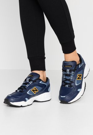 Voeten met marineblauwe en lichtblauwe New Balance sneakers met geel logo, gecombineerd met zwarte strakke broek tegen een effen achtergrond.