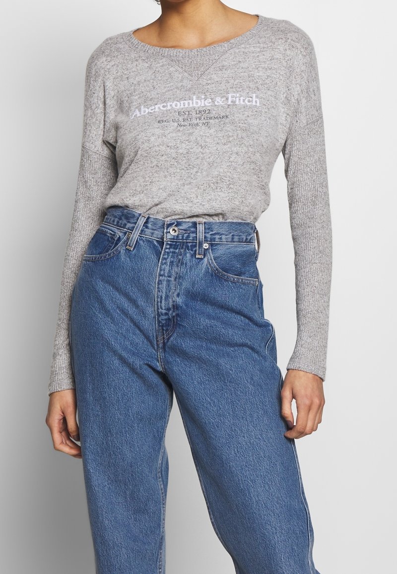 Personne portant un haut à manches longues gris clair Abercrombie & Fitch rentré dans un jean bleu taille haute avec fermeture par bouton et zip.