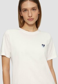 Witte katoenen t-shirt met korte mouwen, voorzien van een klein hartontwerp in tinten van blauw op de linkerborst. Rond decolleté.