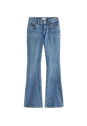 Ung pige står iført en hvid kortærmet skjorte, blå jeans og hvide sneakers mod en ensartet hvid baggrund.