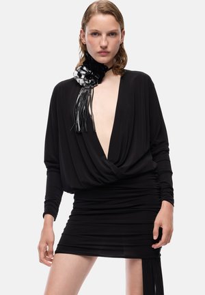 Mujer con un mini vestido negro de escote en V profundo, mangas largas y un gran accesorio de flor negra en el cuello, posando frente a un fondo liso.