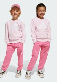 Sweatshirts roses et jogging assortis avec logo Adidas blanc, dotés d'une coupe décontractée et de poignets côtelés, portés avec des baskets noires et blanches.