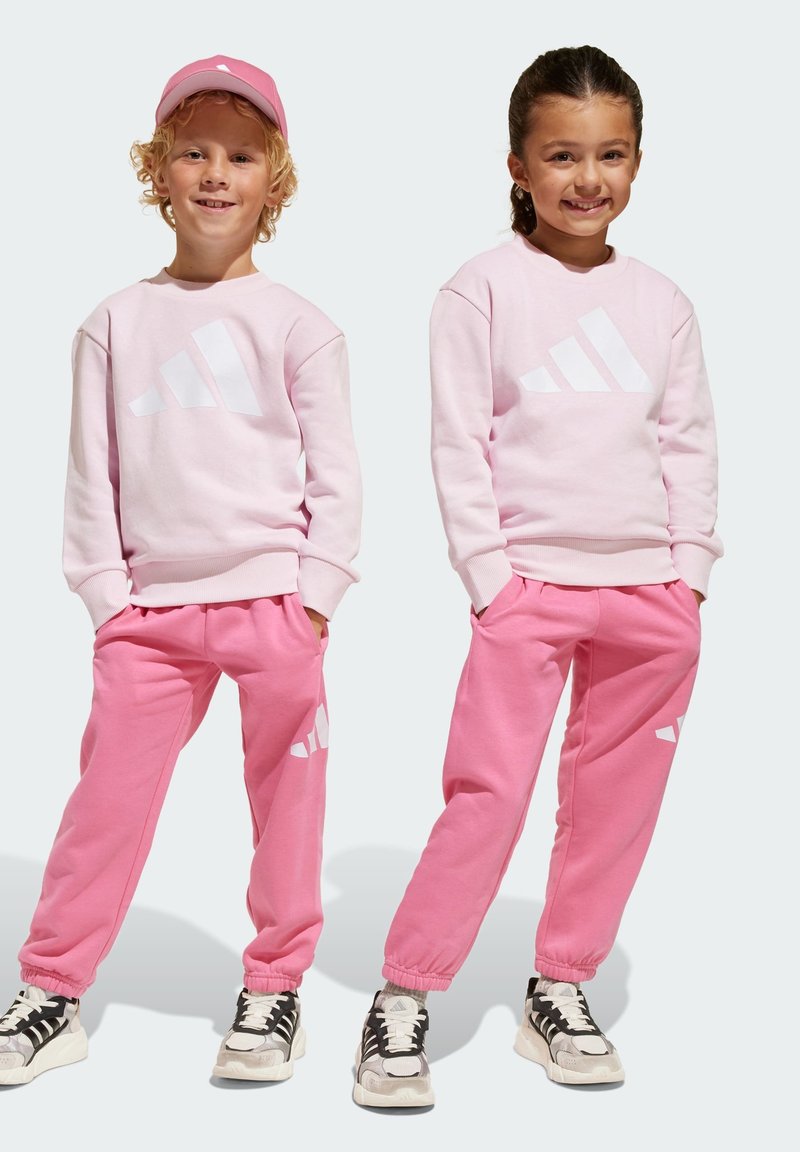 Sweatshirts roses et jogging assortis avec logo Adidas blanc, dotés d'une coupe décontractée et de poignets côtelés, portés avec des baskets noires et blanches.
