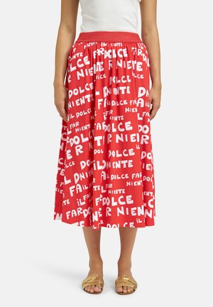 Frau trägt einen roten Midirock mit weiß verstreutem Text, ein weißes ärmelloses Oberteil und goldene Sandalen mit offenen Zehen, steht vor einem schlichten Hintergrund.