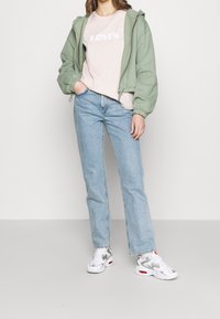 Grön huva bomberjacka, ljusrosa Levi's t-shirt, blå raka jeans och vita sneakers med grå detaljer. Avslappnad outfit visad.