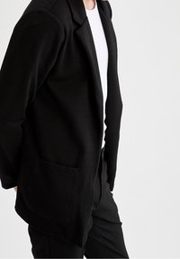 Svart blazer i texturerat tyg med ett herringbone-mönster, med en enda framficka och notchade slag. Stylad över en vit topp.