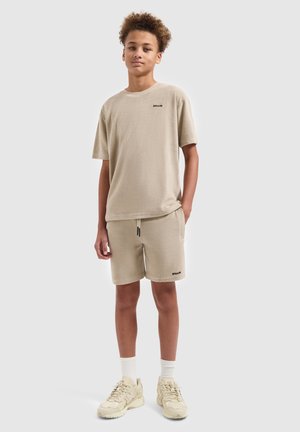 Tiener draagt beige terrydoek T-shirt met korte mouwen en bijpassende korte broek met sneakers, staand tegen een effen lichte achtergrond.