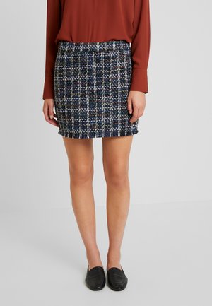Vrouw die een donkerblauwe tweed minirok met wit ruitpatroon draagt, een roestkleurige blouse met lange mouwen en zwarte instapschoenen, staand op een grijze vloer.