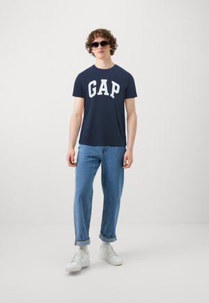Tricou navy cu litere albe "GAP", asortat cu blugi de culoare bleu deschis și adidași albi. Modelul poartă ochelari de soare închiși la culoare și stă în fața unui fundal simplu.