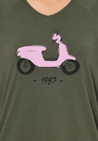 Camiseta verde oliva con un gráfico de scooter rosa y el texto "1987" debajo. Tejido suave y ligero con un corte relajado y diseño de escote en forma de V.