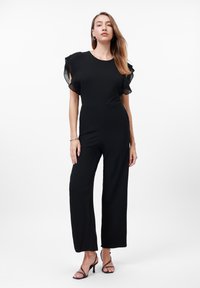 Tuta nera con busto aderente, maniche a ruffle plissettate e pantaloni a gamba larga. Tessuto liscio con una leggera lucentezza, design minimalista.