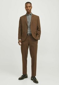 Costume marron avec une coupe ajustée, associé à une chemise grise à motifs. Une ceinture et des chaussures noires complètent le look. Tissu lisse, design moderne.