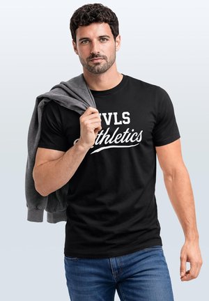 Man with dark curly hair wearing black "DVLS Athletics" t-shirt and blue jeans, holding gray sweatshirt over shoulder.