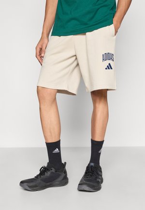 Pantalones cortos beige de corte relajado, con el logo de "Adidas" en azul marino en la pierna izquierda. Combinados con zapatillas deportivas negras y calcetines oscuros.