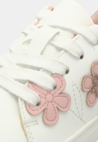 Λευκό αθλητικό παπούτσι με ροζ κεντήματα λουλουδιών, κορδόνια με υφή και λεία επιφάνεια. Στοιχεία διακόσμησης περιλαμβάνουν ραμμένα σχέδια λουλουδιών και διακριτικές λεπτομέρειες.