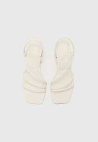 Sandales à talons blancs avec un bout carré, présentant de multiples fines brides croisées et une finition lisse et brillante. Logo doré embossé sur la semelle intérieure.