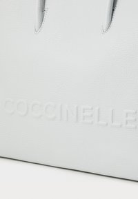 Svijetlosiva kožna torba s teksturom s ugraviranim Coccinelle logotipom na prednjoj strani. Dvostruki gornji ručkA za jednostavno nošenje.