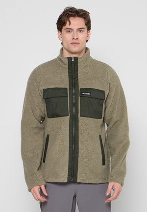 Giovane uomo con giacca in pile beige e verde scuro con cerniera frontale e due tasche con patta sul petto, in piedi davanti a uno sfondo semplice.