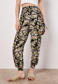 Pantalons à motif feuille noir et or avec une taille élastique et des chevilles resserrées, en tissu léger. Associés à des sandales à glisser beiges.