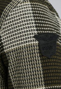 Chemise en tissu tricoté avec un motif texturé en vert olive et crème. Présente un patch tissé noir avec un aigle et "PME 1649-A".